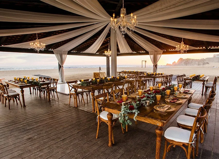Beachside outdoor banquet setup with long wooden tables, chairs, floral centerpieces, chandeliers, and draped fabric, overlooking the ocean at sunset.