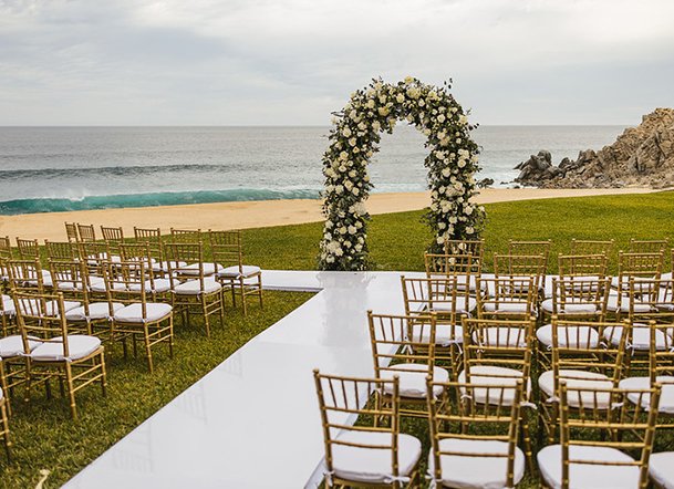 Outdoor wedding setup on a grassy area near the beach with rows of gold chairs, a white aisle, and a floral arch.