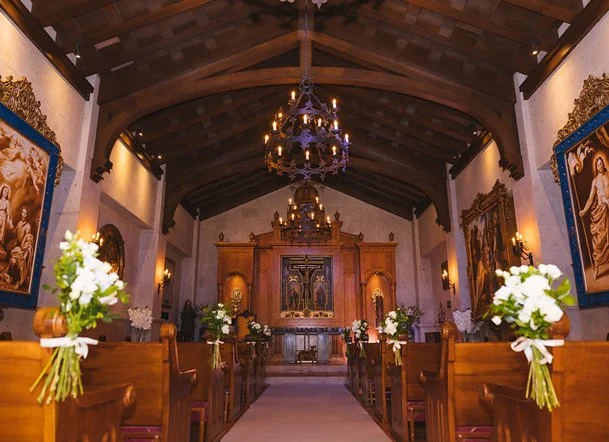 Interior of a church with wooden pews decorated with white flowers, an altar with religious artwork at the front, and chandeliers hanging from a dark wooden ceiling.