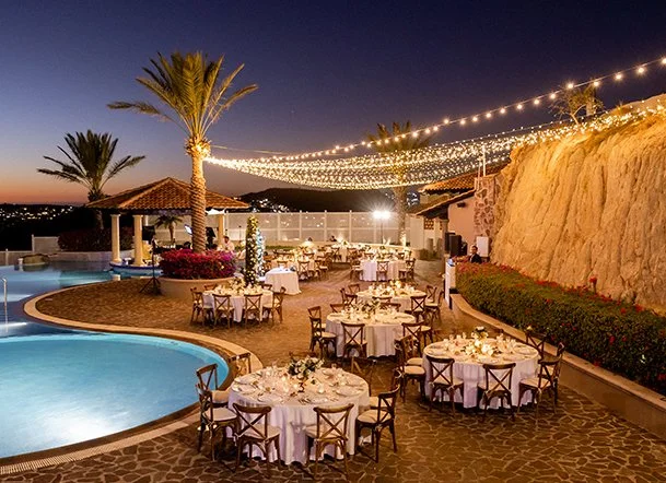 Outdoor evening party with round tables set with white tablecloths and chairs, a pool, palm trees, string lights, and a hillside backdrop.