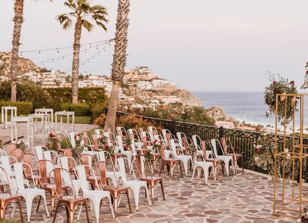 Outdoor wedding ceremony setup with white, pink, and copper-colored chairs on a stone patio, overlooking the ocean with palm trees, lights, and hillside buildings in the background.