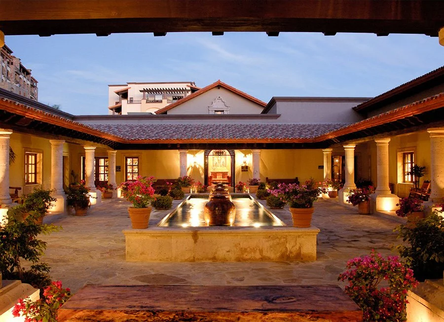 A sunny courtyard with yellow walls, columns, and a central water fountain surrounded by potted pink and purple flowers, with buildings in the background.