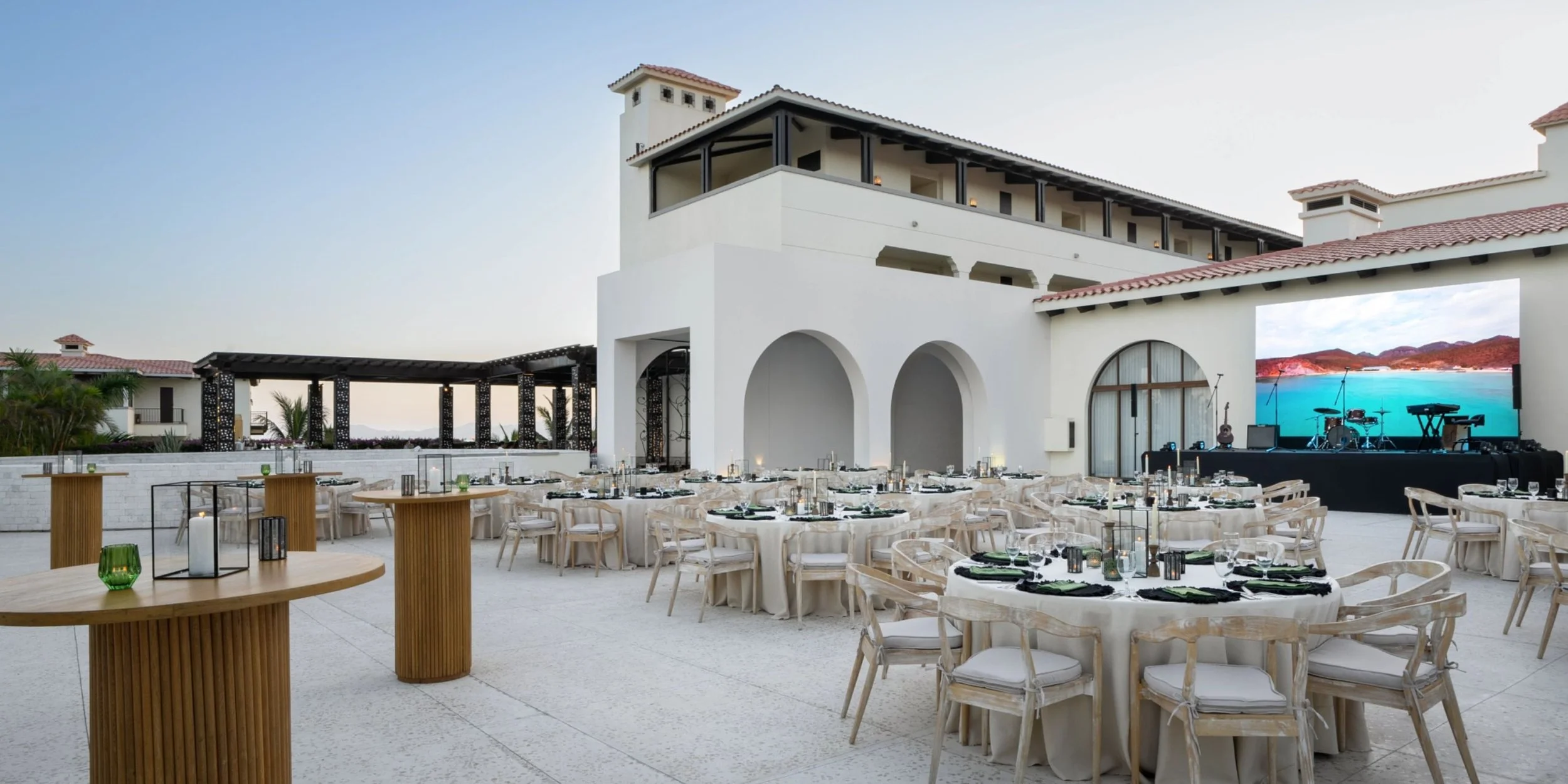 Outdoor event setup with round tables, chairs, and stage at a waterfront venue with a large screen and musical instruments.