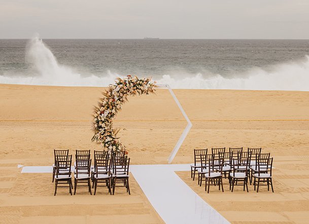 Wedding ceremony setup on a sandy beach with chairs arranged in front of a floral arch and ocean waves crashing in the background.