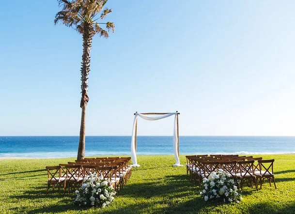 Beachside wedding setup with wooden chairs, floral arrangements, and a fabric-draped arch under a palm tree, overlooking the ocean.