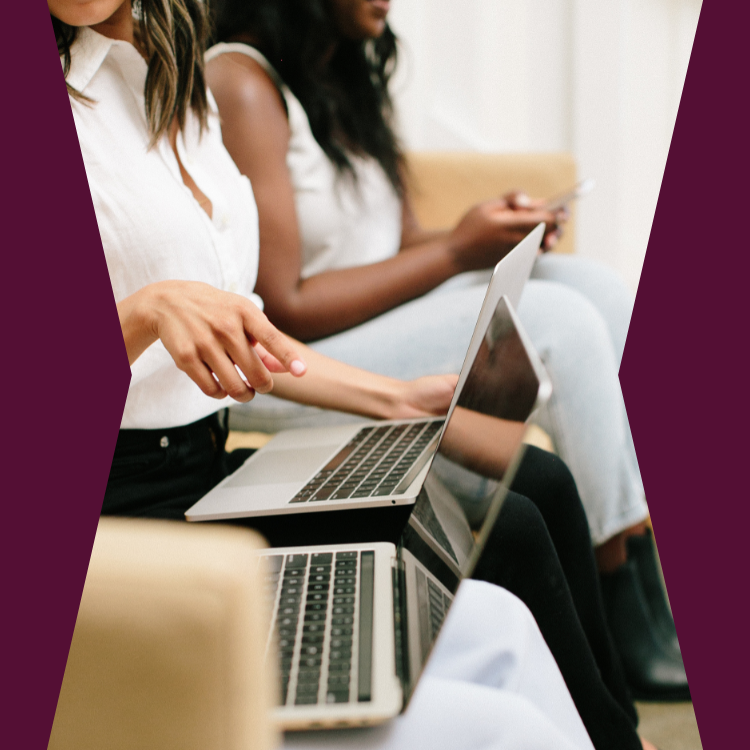 Two women sitting on a couch with laptops and a phone, working or browsing.