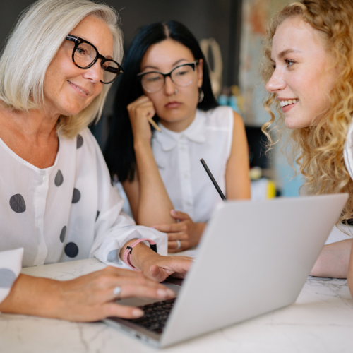 Three women, two younger and one older, sitting around a laptop, engaged in a discussion.