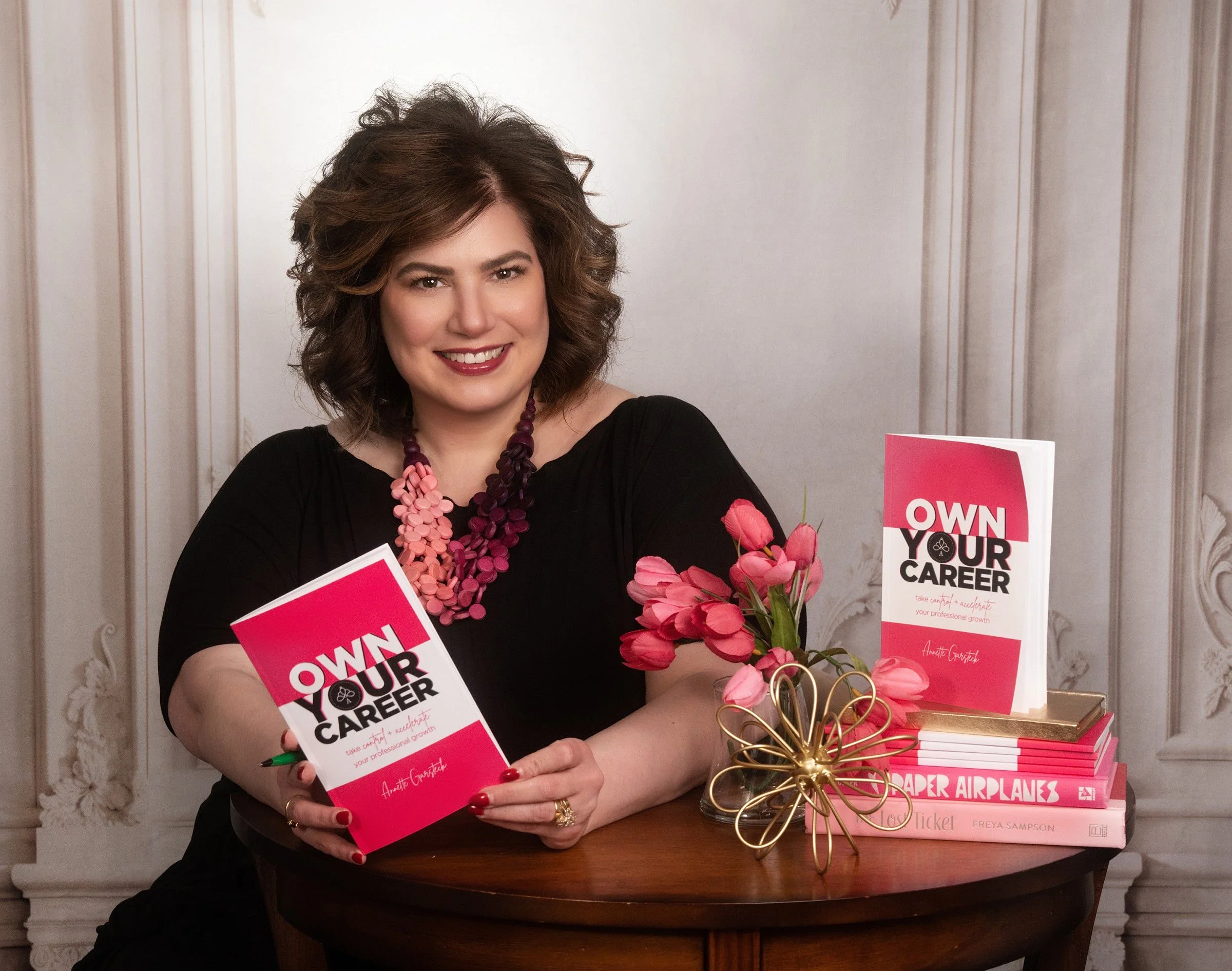 Annette Garsteck is wearing a black top and a pink and purple floral necklace, sitting at a table with copies of her books titled 'Own Your Career' and a bouquet of pink flowers, smiling at the camera.