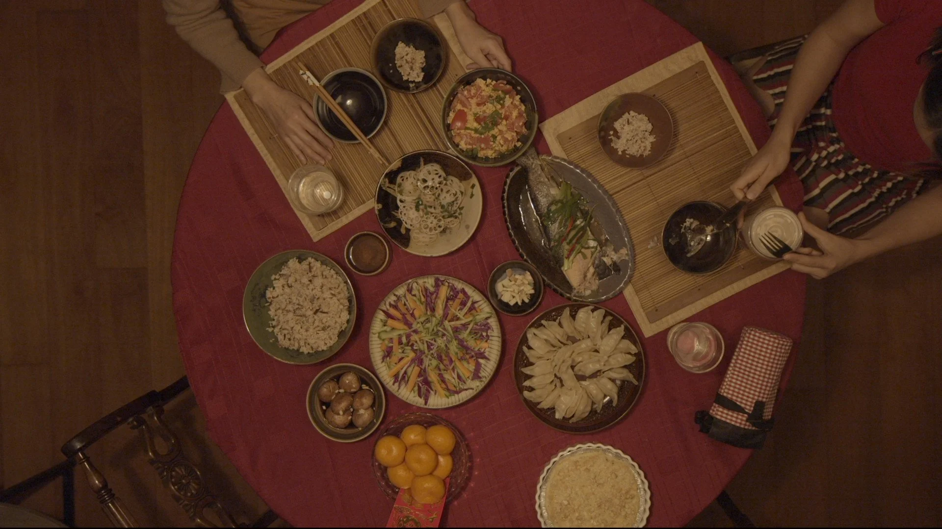 An image from the perspective of above of a dinner table covered in a red table cloth and many plates of Chinese food. A set of hands at one of the placements and the arms of girl dishing up a meal are seen.   
