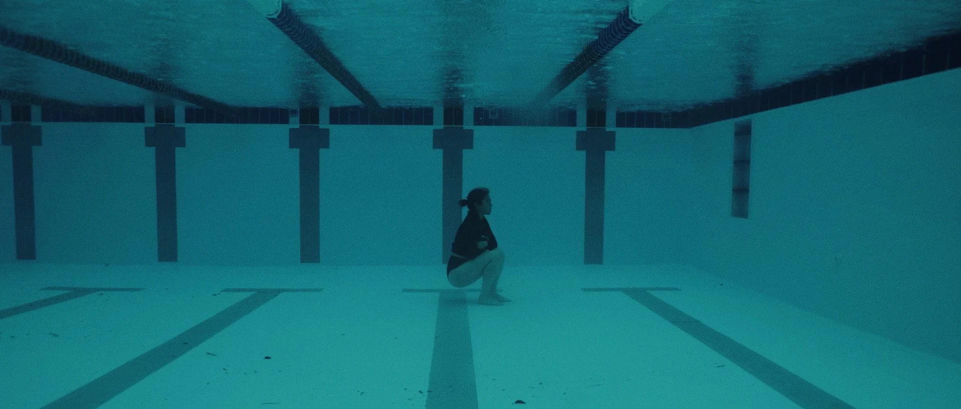A younf Asian woman in a black long sleeve bathing suit is squatting at the bottom of a lap pool under water.