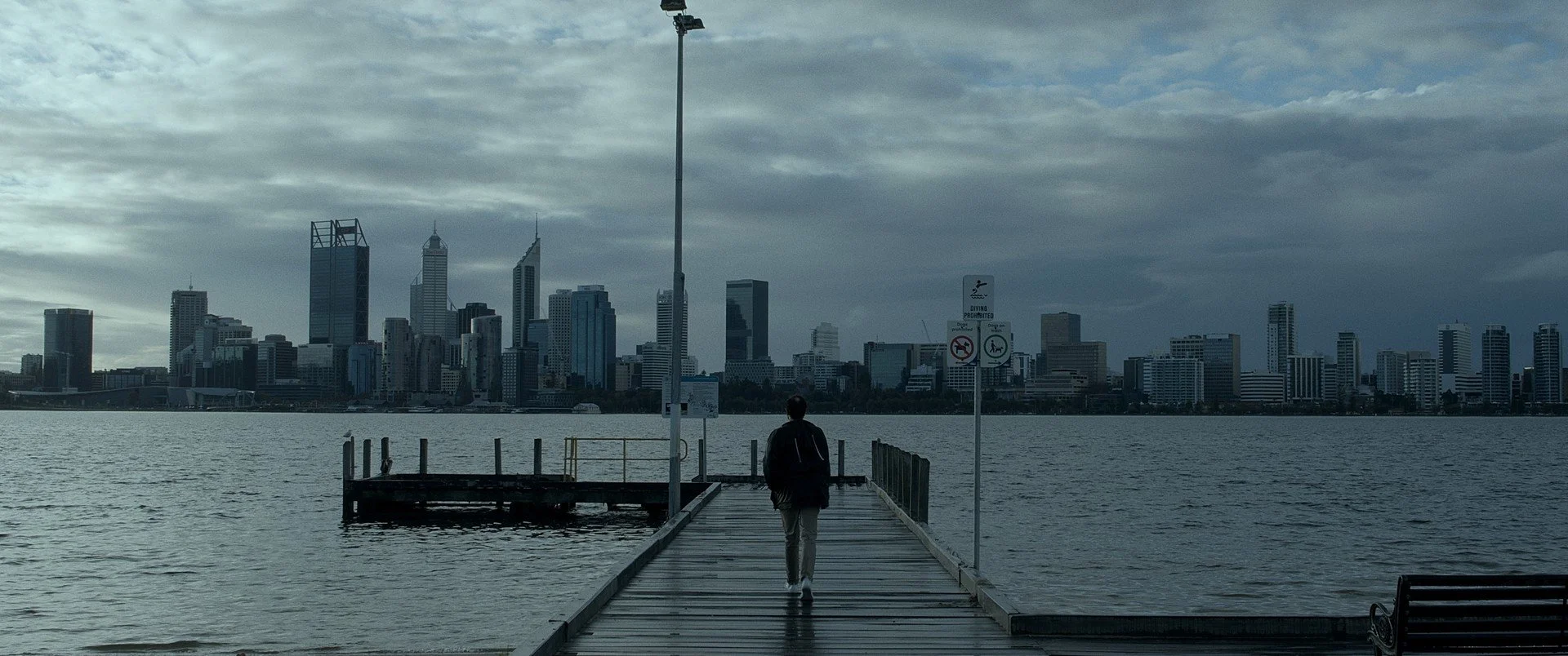An image of a city skyline on a grey cloudy day. In the foreground is a pier with a young man walking down it, his back facing us.