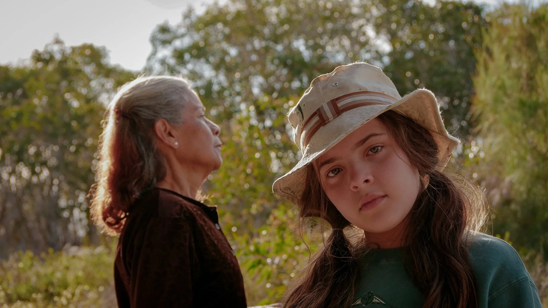 An image of a girl from the shoulders up wearing a green top and beige fisherman's hat. Behind her an older woman in a brown cardigan is standing in profile. There are trees in the background.