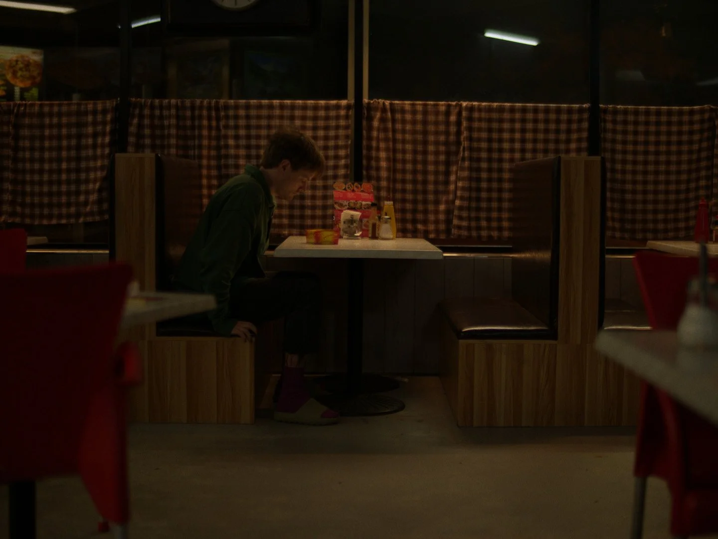 A man in his 20s is sitting a booth at a dimly lit café alone looking down at some fat food on the table.