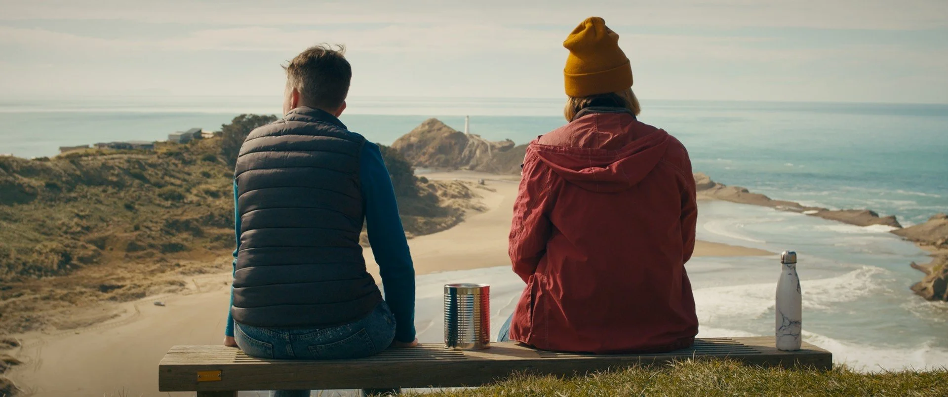 Two white women are sitting on a wood bench with backs facing us. One is wearing a black puffer vest with a long-sleeved green top underneath and jeans. The other is wearing a mustard beanie and red rain jacket. They are looking over a coastline. 