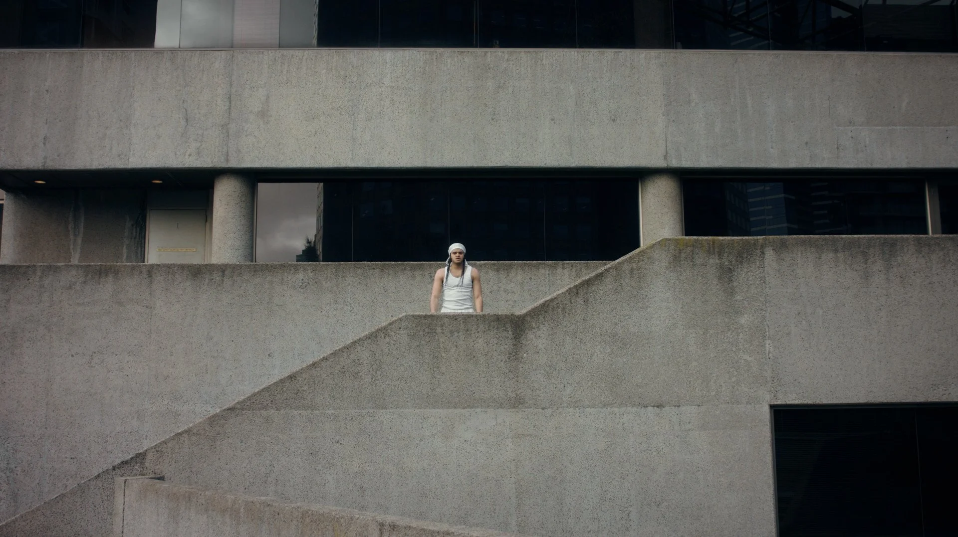 A young man in a white singlet and head bandana stands on a large cement staircase surrounded by a cement building. 