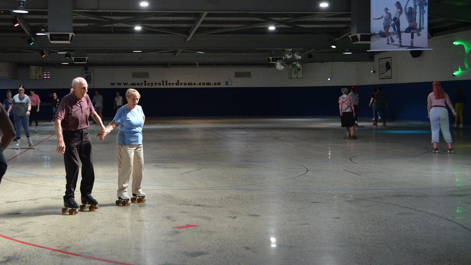 An elderly white couple are holding hands as they roller skate around a rink. Ther are also other people rolling skating in the background.