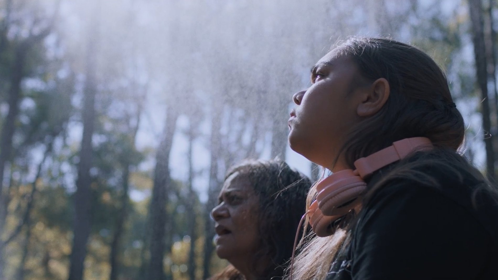 A young Indigenous girl and an older Indigenous woman are looking off to the side of the frame into the forest of trees that surround them.