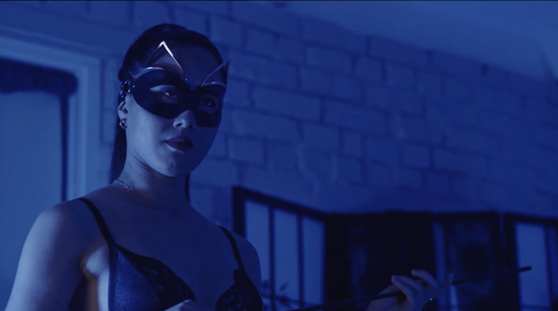 An image of a woman from the chest up. She is wearing a cat-like black mask across her eyes and a black bra. She is in a dimly lit brick room with a dressing screening in the background.