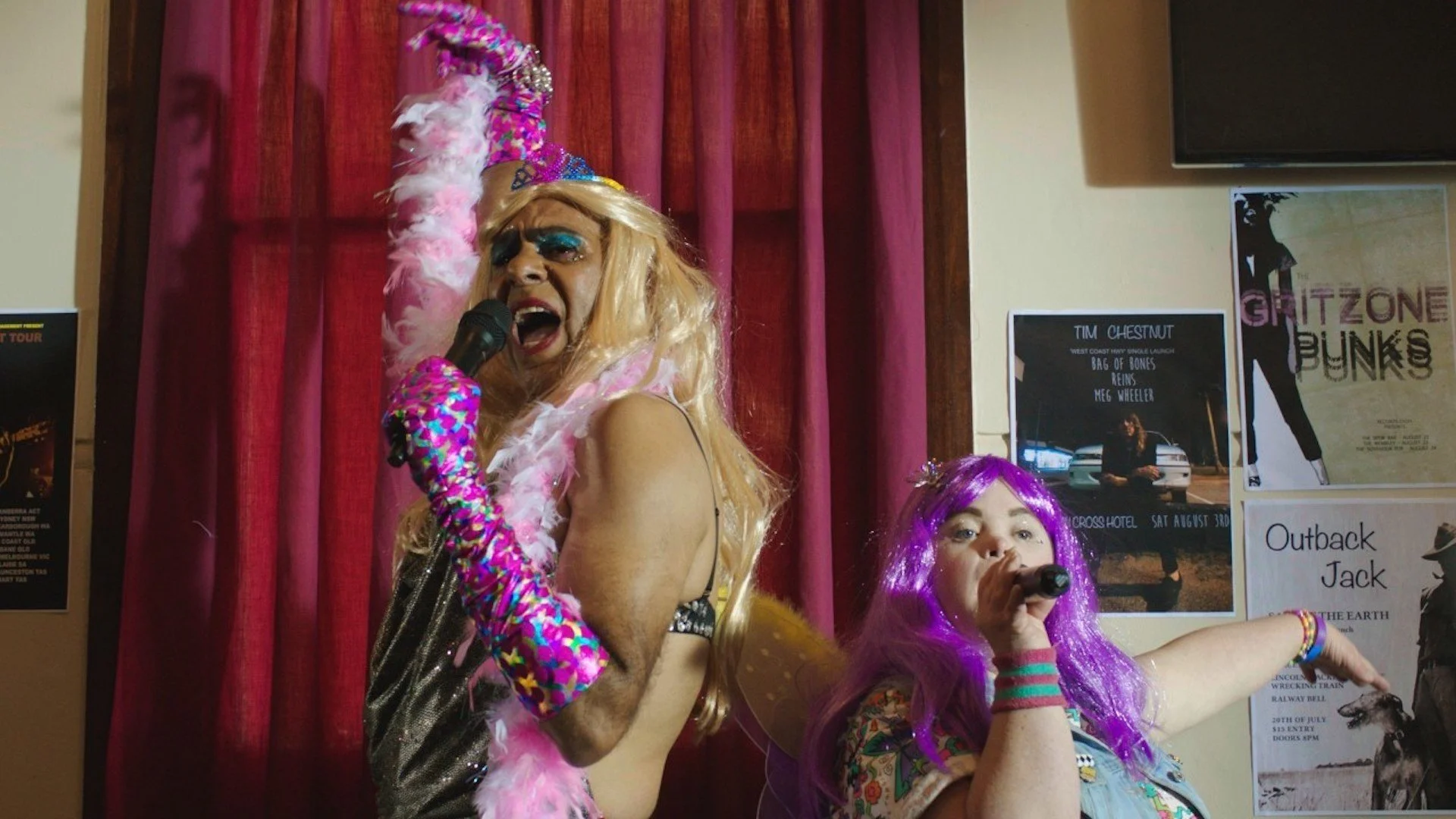 A drag queen dressed in pink and young woman with Down Syndrome wearing a purple wig are holding microphones and singing kareoke.