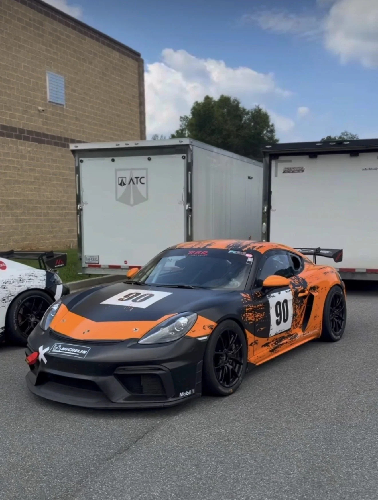 A Porsche race car painted in black and orange with the number 90 on the hood and side, parked on a street near a brick building with two large trailers in the background.