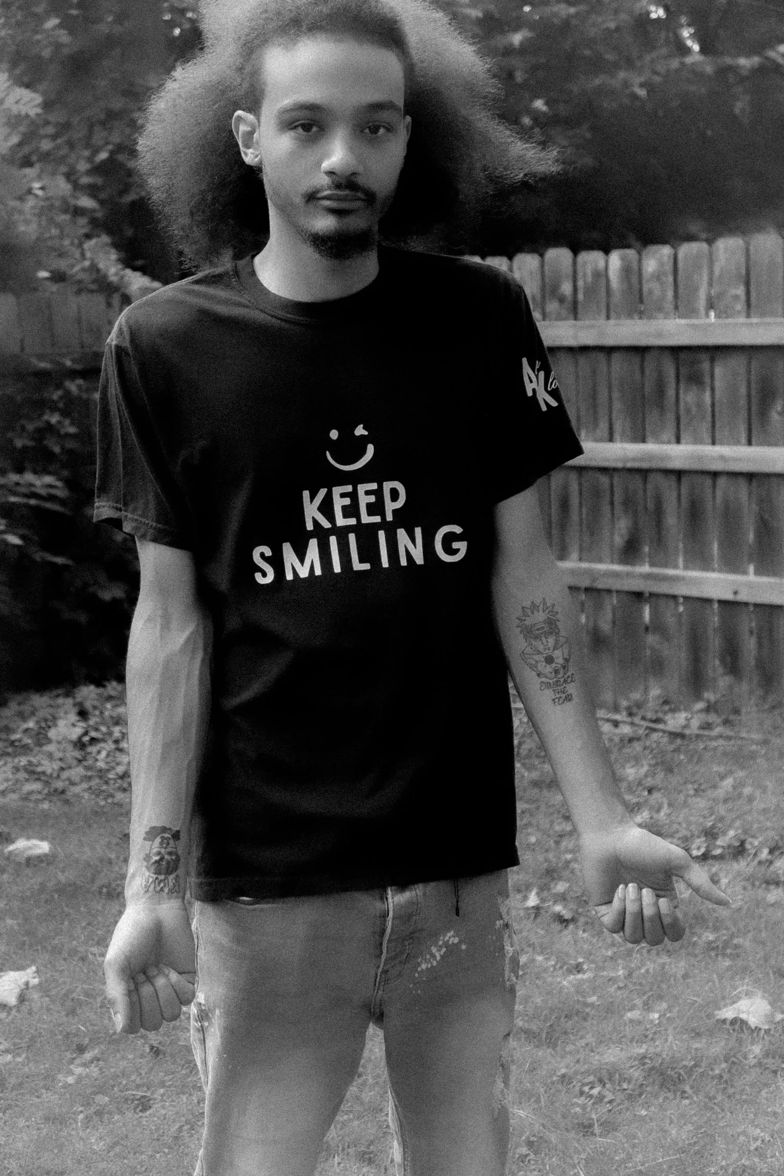 A young man with long, curly hair wearing a t-shirt that says 'Keep Smiling' and has a smiley face, standing outdoors with a wooden fence and trees in the background.