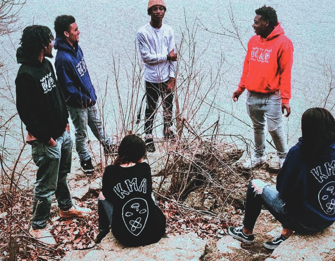 Seven young people gathered outdoors on a rocky area near a body of water, engaged in conversation. Some are standing, while others are sitting on the ground or rocks. They are dressed casually in hoodies, jackets, and sneakers, with two of them wear