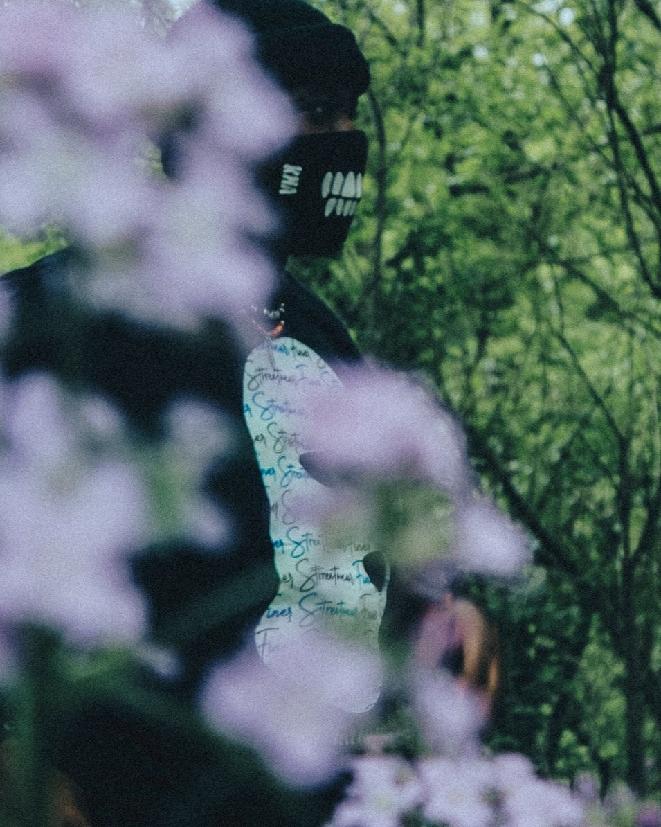 Person wearing a black mask with white teeth design and black hoodie, standing among trees and pink flowers, with a blurry background.