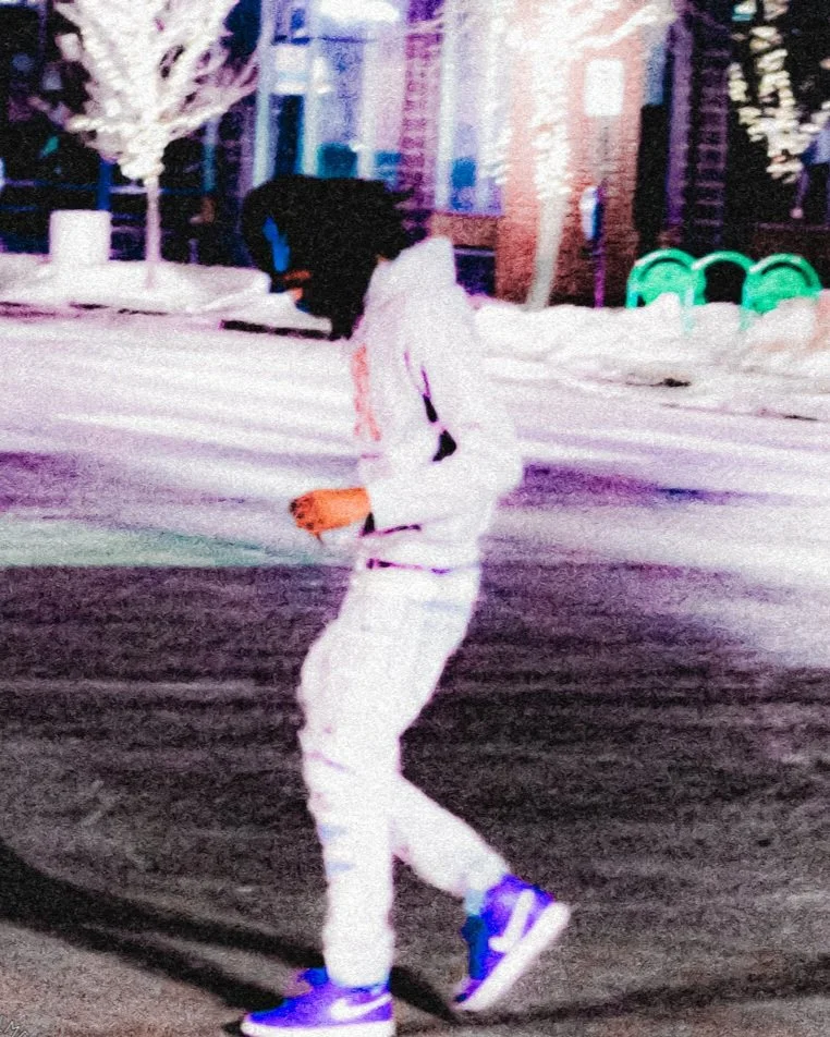 Person walking outdoors at night near buildings and trees, wearing a white hoodie, white pants, and blue shoes.