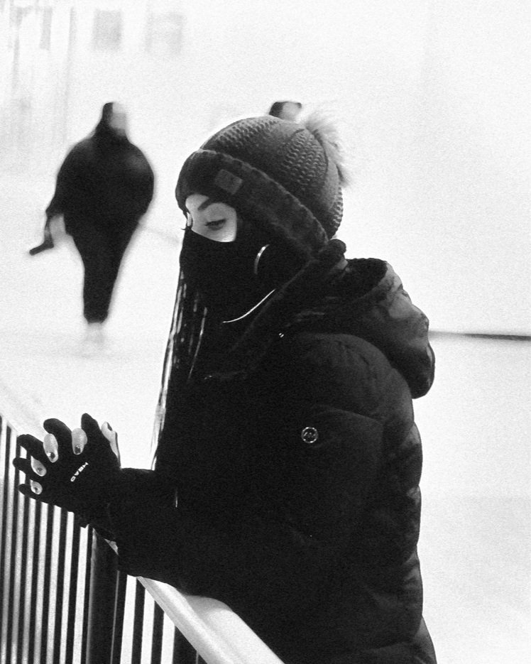 Person wearing a knitted hat, face mask, and gloves, standing by a railing in an indoor setting, with a woman walking away in the background.