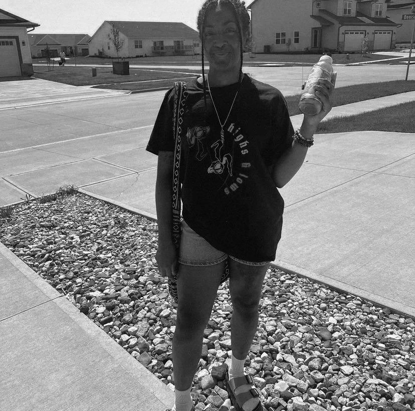 A woman standing outdoors on a sunny day, holding a drink bottle, wearing a black t-shirt, shorts, and sandals, with houses and a driveway in the background.
