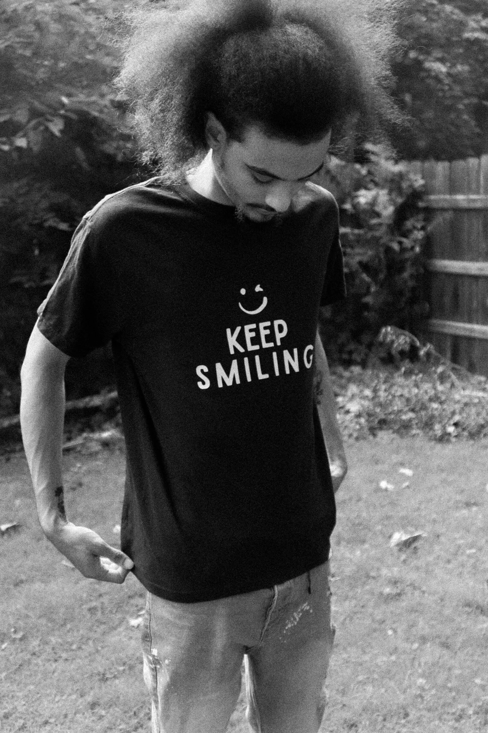 A person with curly hair wearing a black t-shirt that says 'Keep Smiling' with a smiley face and jeans, standing outdoors in a garden.