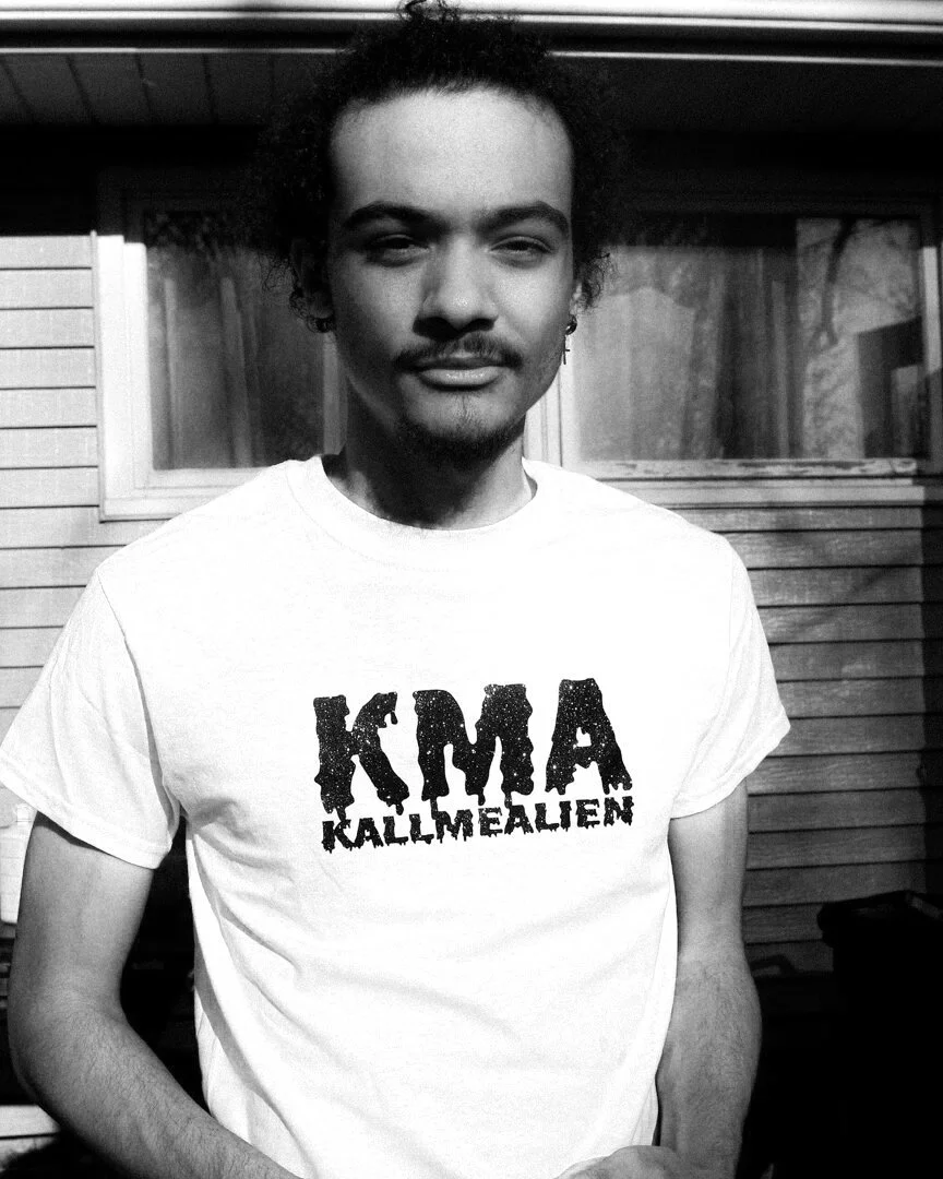 A young man with curly hair, a mustache, and beard, wearing a white t-shirt with the words 'KMA KALLMEALEN' printed on it, standing outside in front of a house with a window and wooden siding.