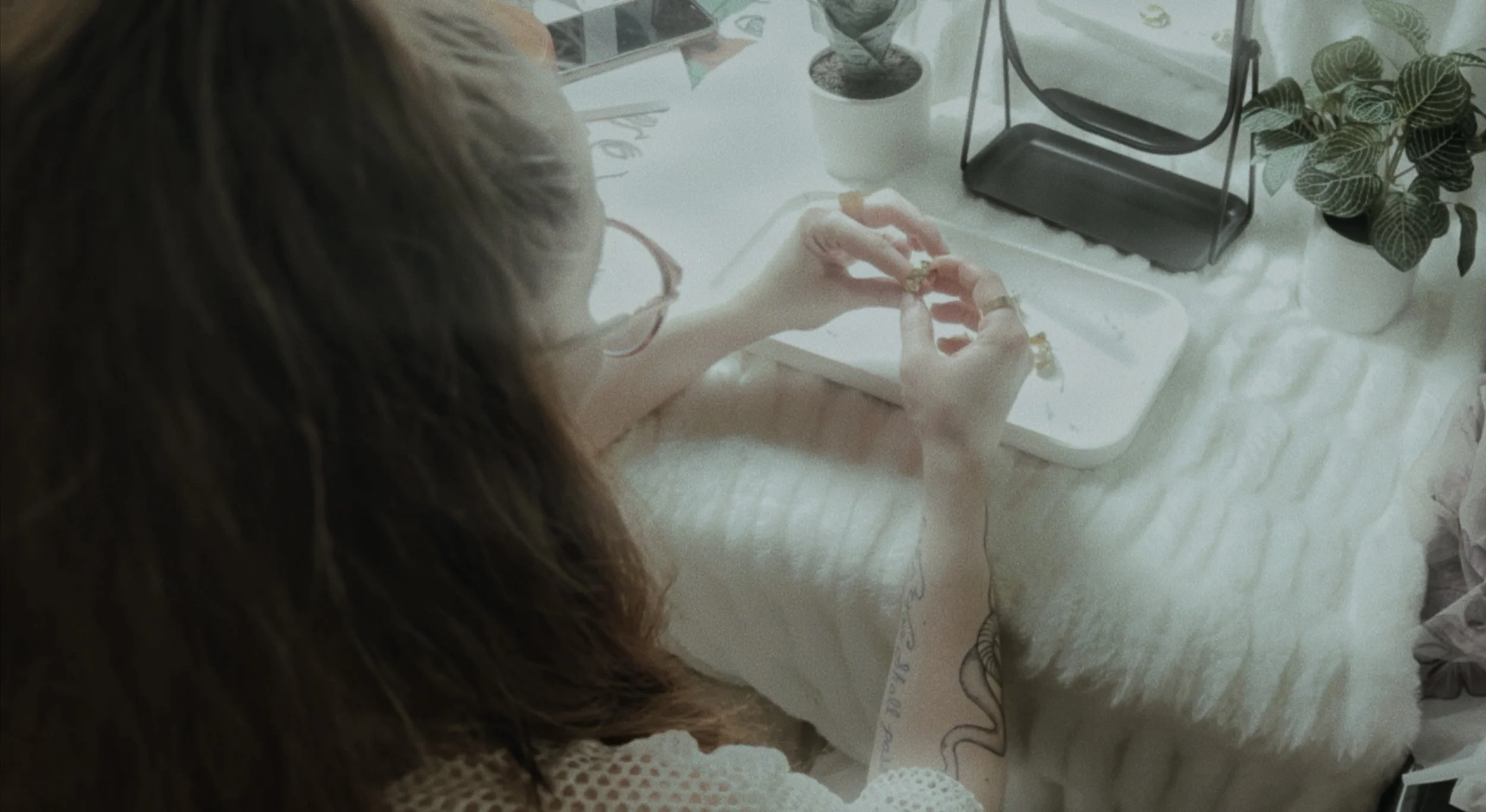 A person with long brown hair wearing glasses, sitting at a table, is holding jewelry and working on it with their hands. There are plants, a tray, and some stationery on the table, which has a fluffy white tablecloth.