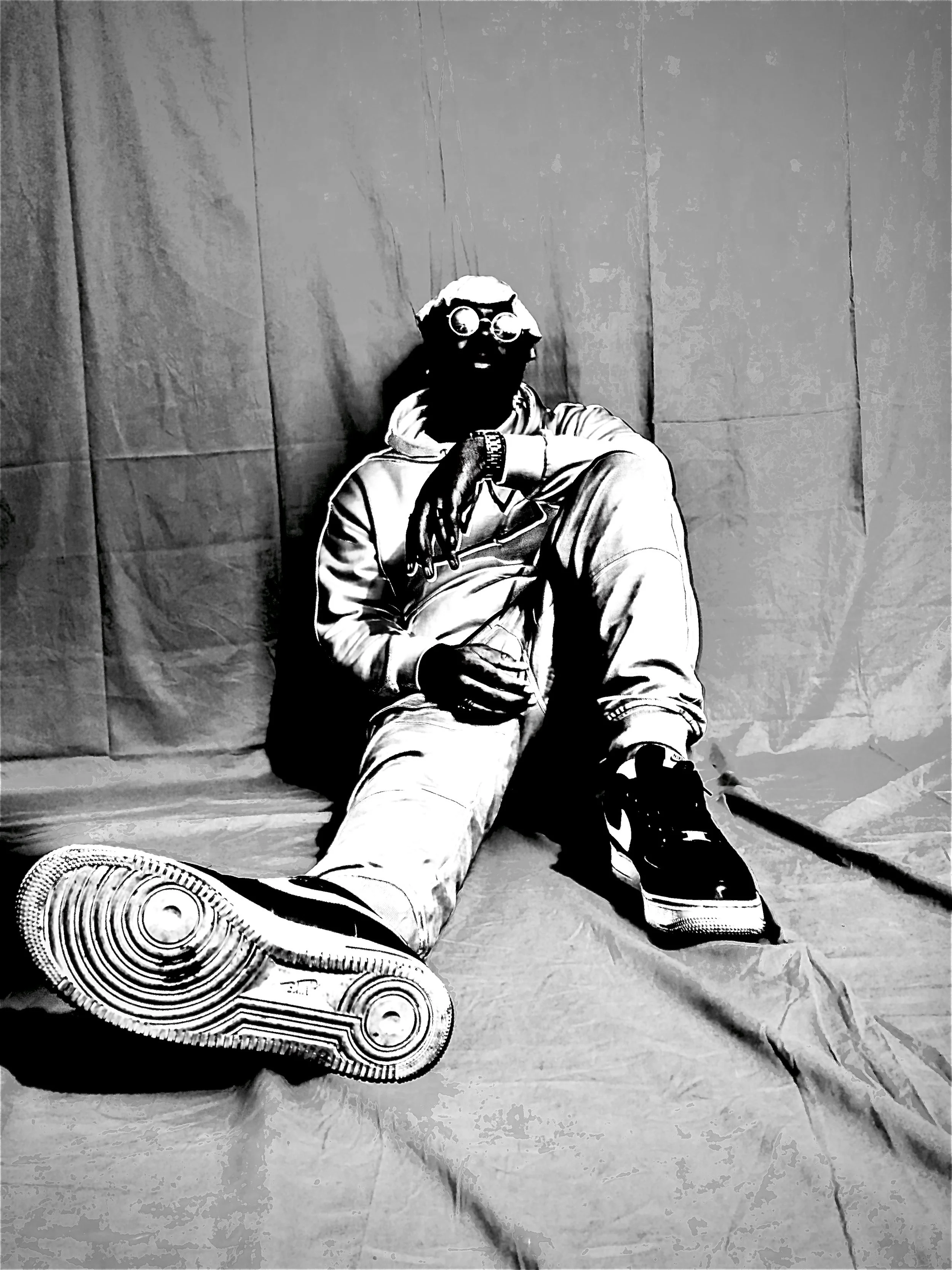 Black and white photo of a person sitting on the floor with a relaxed pose, wearing a hoodie, sunglasses, and sneakers, against a plain backdrop.