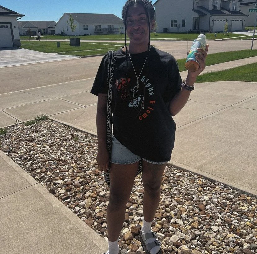 Young woman standing on a sidewalk in a residential neighborhood, holding a bottle in her right hand, wearing a black T-shirt, denim shorts, sandals, and white socks, with a background of houses, grass, and a clear sky.