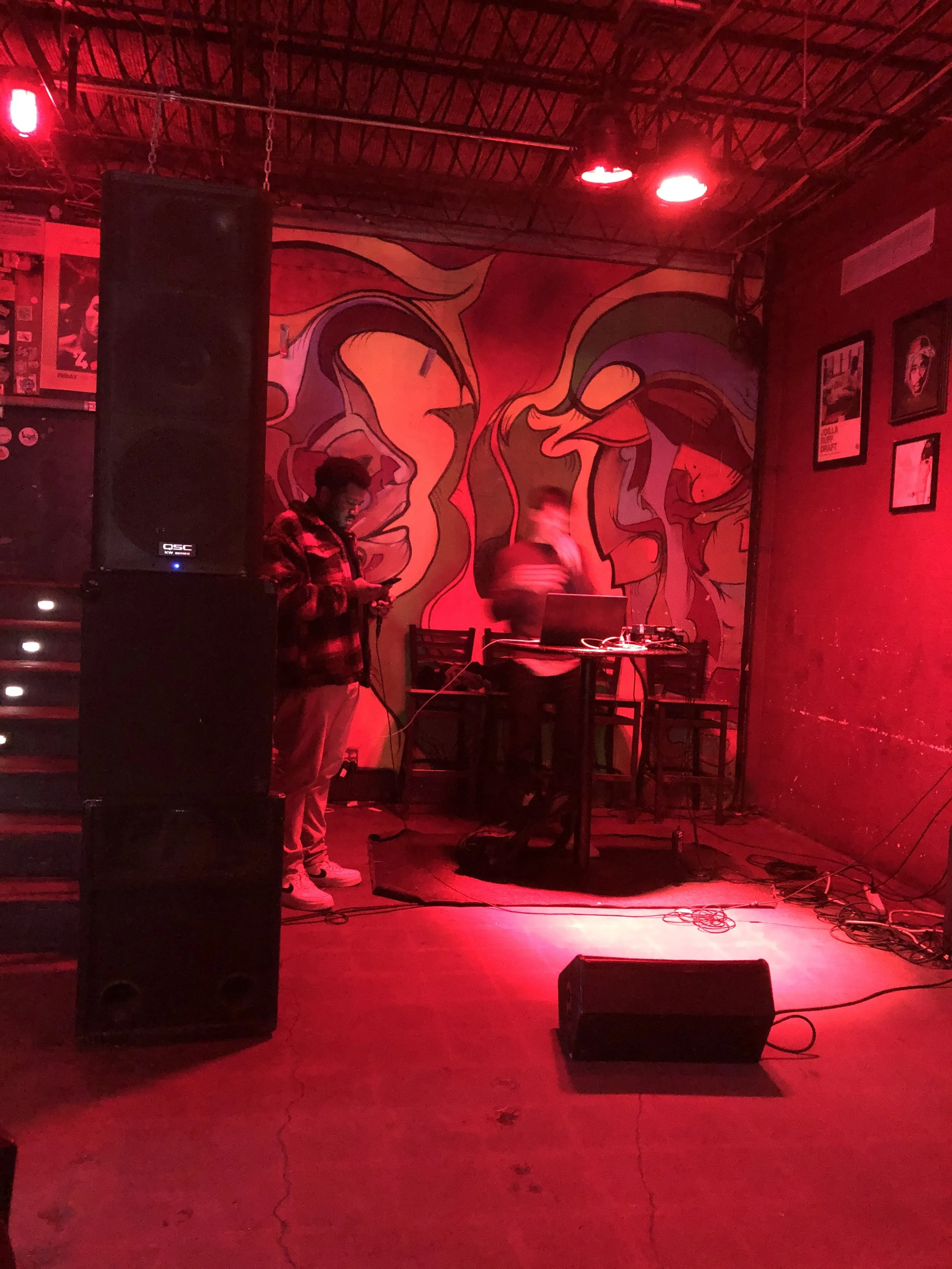 A dimly lit stage with red lighting and a graffiti mural of faces on the back wall. There is a person in a red and black jacket operating DJ equipment at a table, and another person on the left holding a phone, standing near large black speakers.