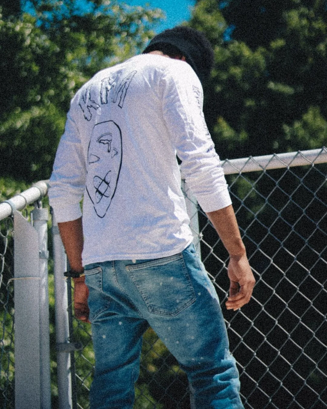 Person in a white long-sleeve shirt with a face sketch and text on the back, leaning on a chain-link fence outdoors on a sunny day.