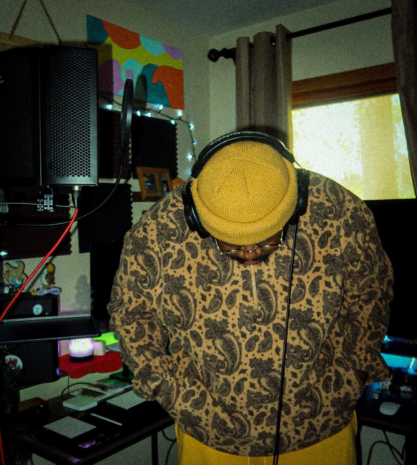 A person wearing a yellow beanie and glasses, with headphones on, is working at a music production setup in a room with colorful wall art, black acoustic panels, and a window with curtains letting in natural light.