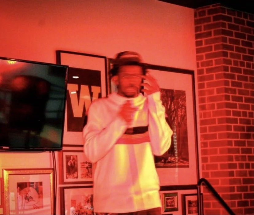 A person with long hair and glasses, wearing a long-sleeve shirt with horizontal stripes, holding a microphone, standing in front of wall art and a television, illuminated by red and yellow lighting.