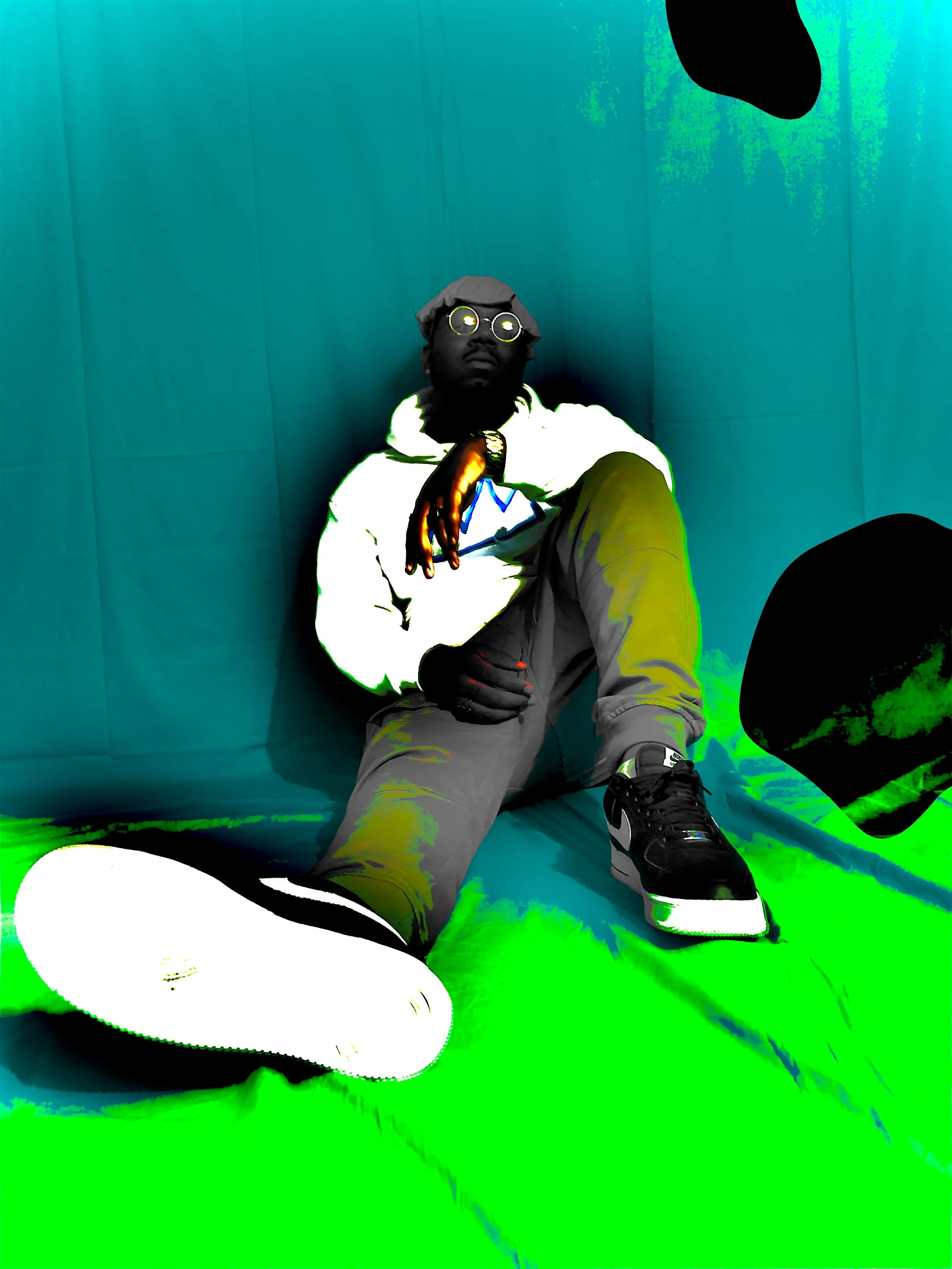 Person sitting on the floor against a green and blue background, wearing glasses, a hoodie, and sneakers, with one leg extended and one arm resting on their knee.