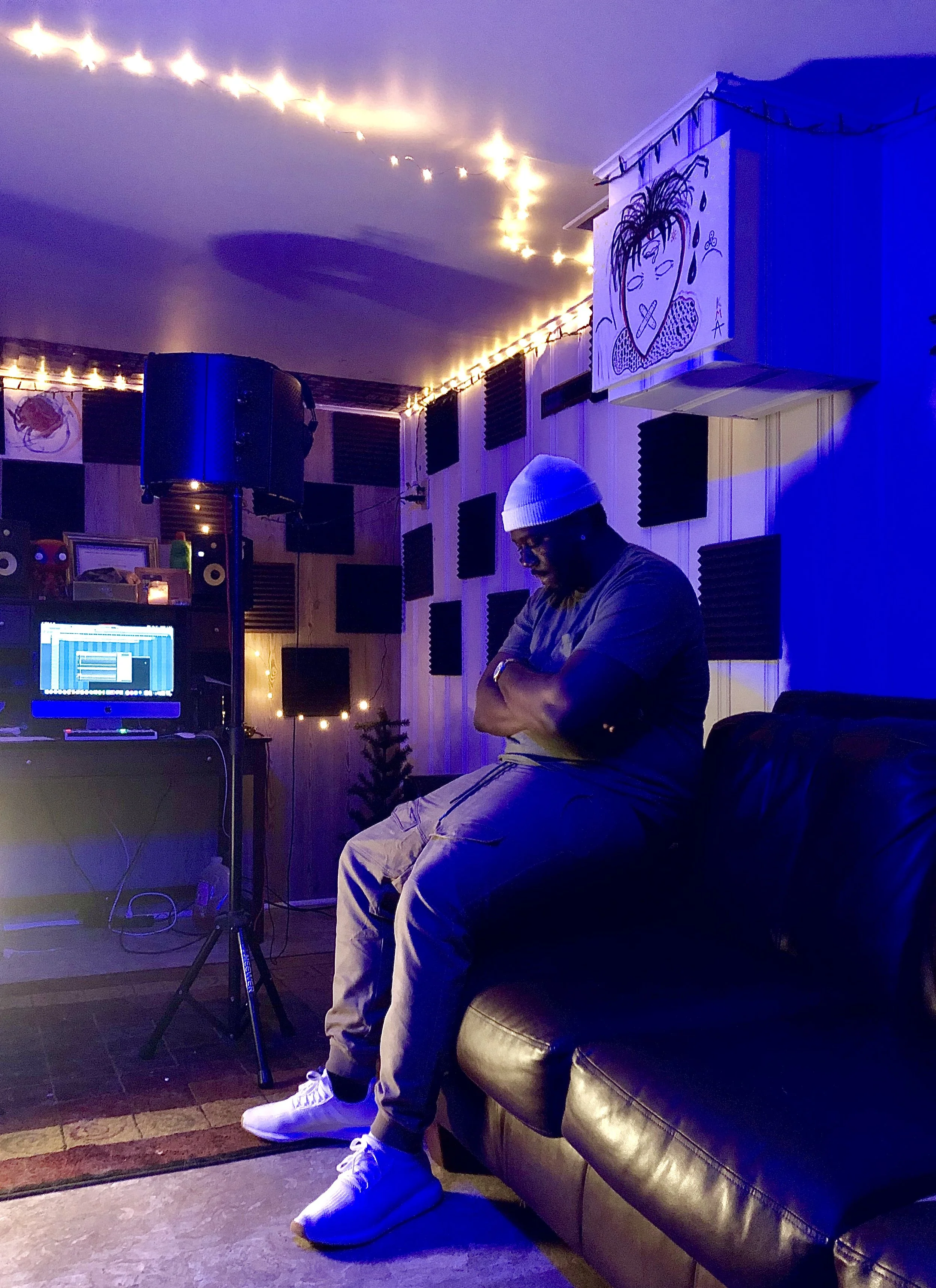 A man in a white beanie and glasses sitting on a black leather sofa with arms crossed, in a dimly lit room with blue lighting, soundproof foam panels on the wall, a drawing of a face with dreadlocks on a hanging box, string lights on the ceiling, a small christmas tree, and a music production setup with a computer and speakers.