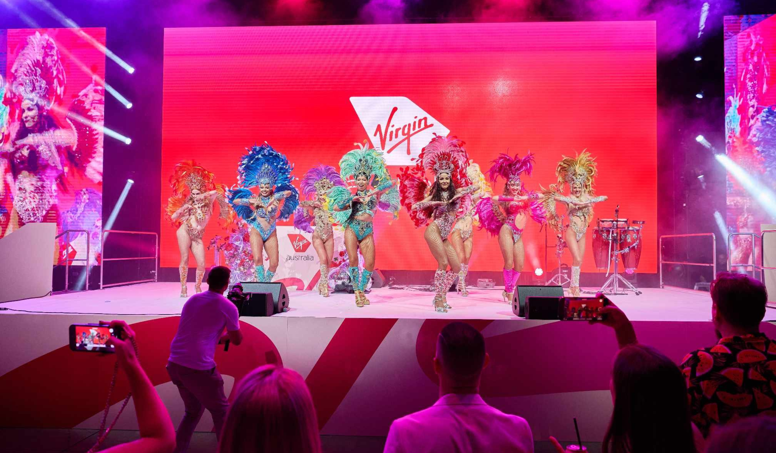 Corporate event crowd facing a large stage with bright pink lighting and oversized LED screens for a live presentation.