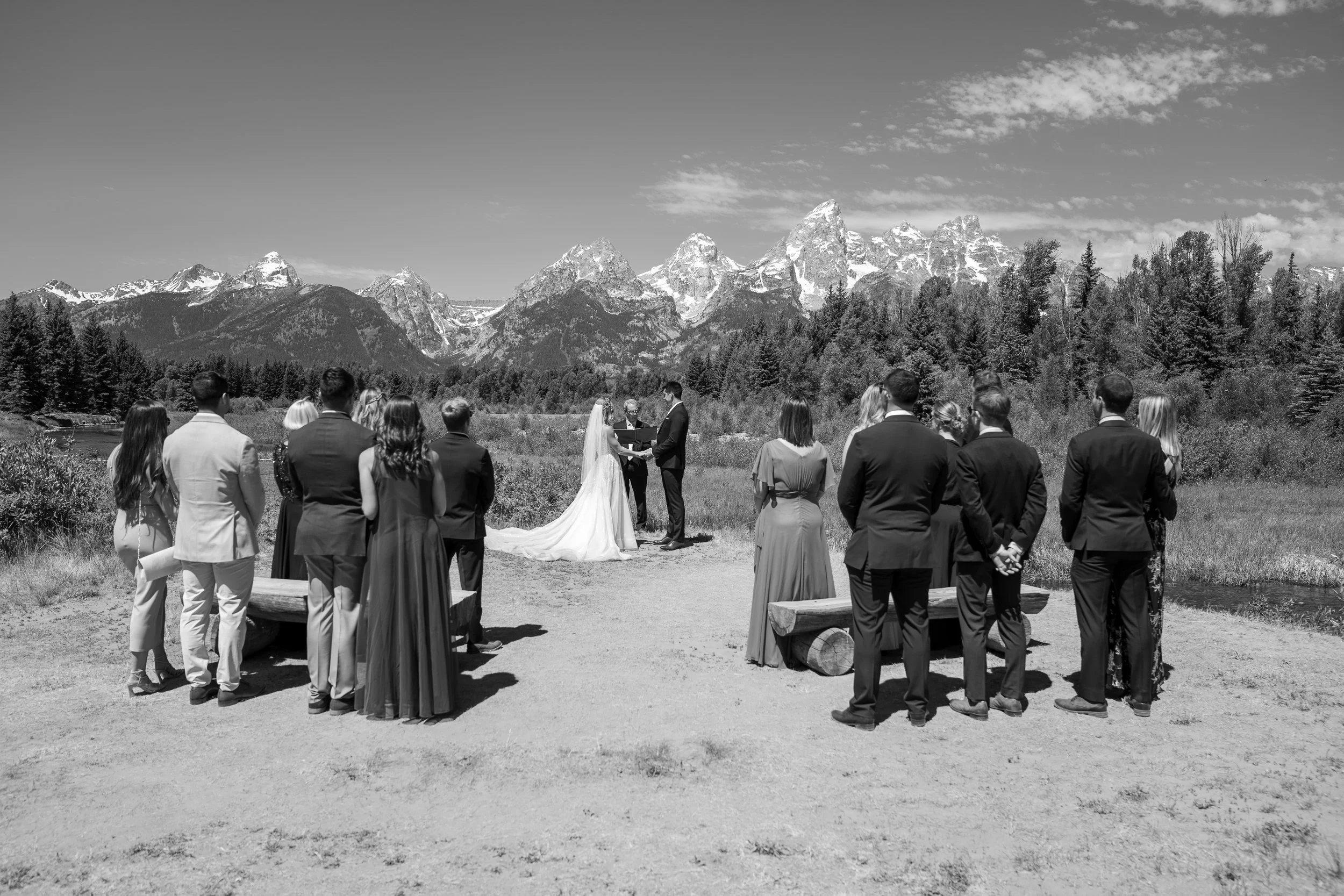 A black-and-white photo of a wedding ceremony outdoors with mountains in the background, featuring a bride and groom exchanging vows before an officiant, surrounded by guests.