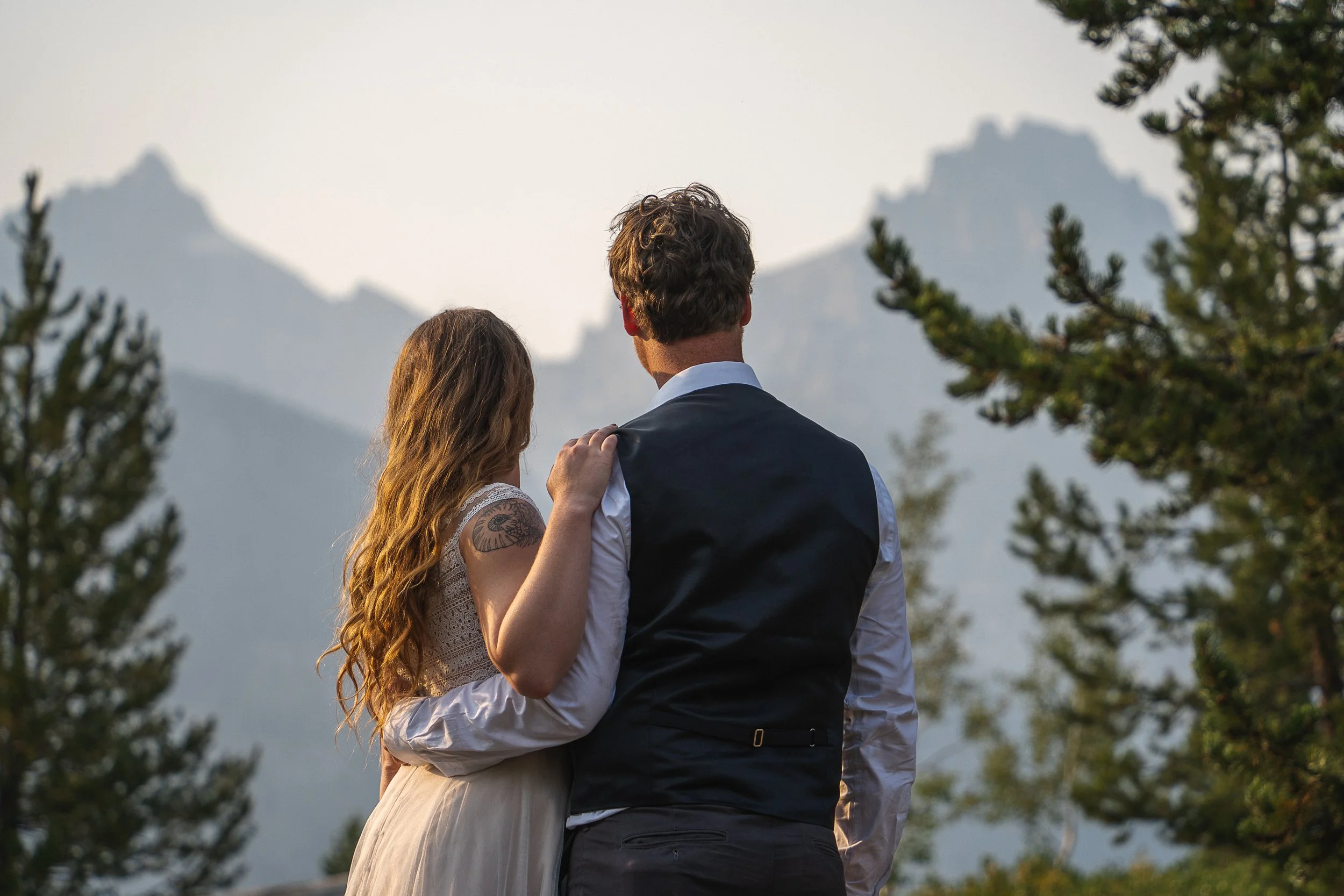 A couple stands outdoors, with mountains and trees in the background, facing away from the camera. The woman has long, wavy hair and a tattoo on her upper arm, resting her hand on the man's shoulder. The man has curly hair and is dressed in a white shirt and dark vest.