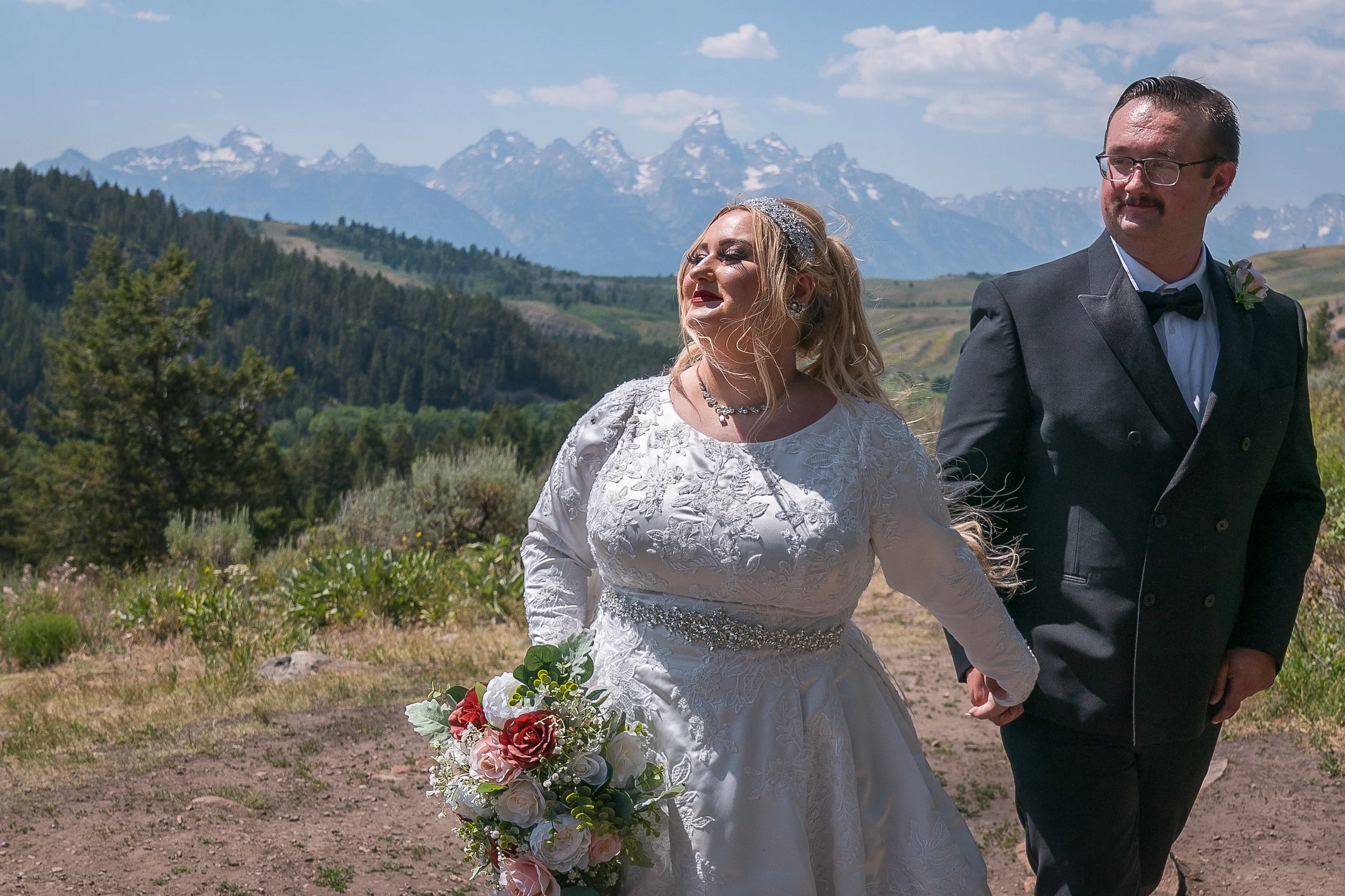 A bride and groom holding hands outdoors with mountains in the background, the bride holding a bouquet of red and white flowers, both dressed in wedding attire.