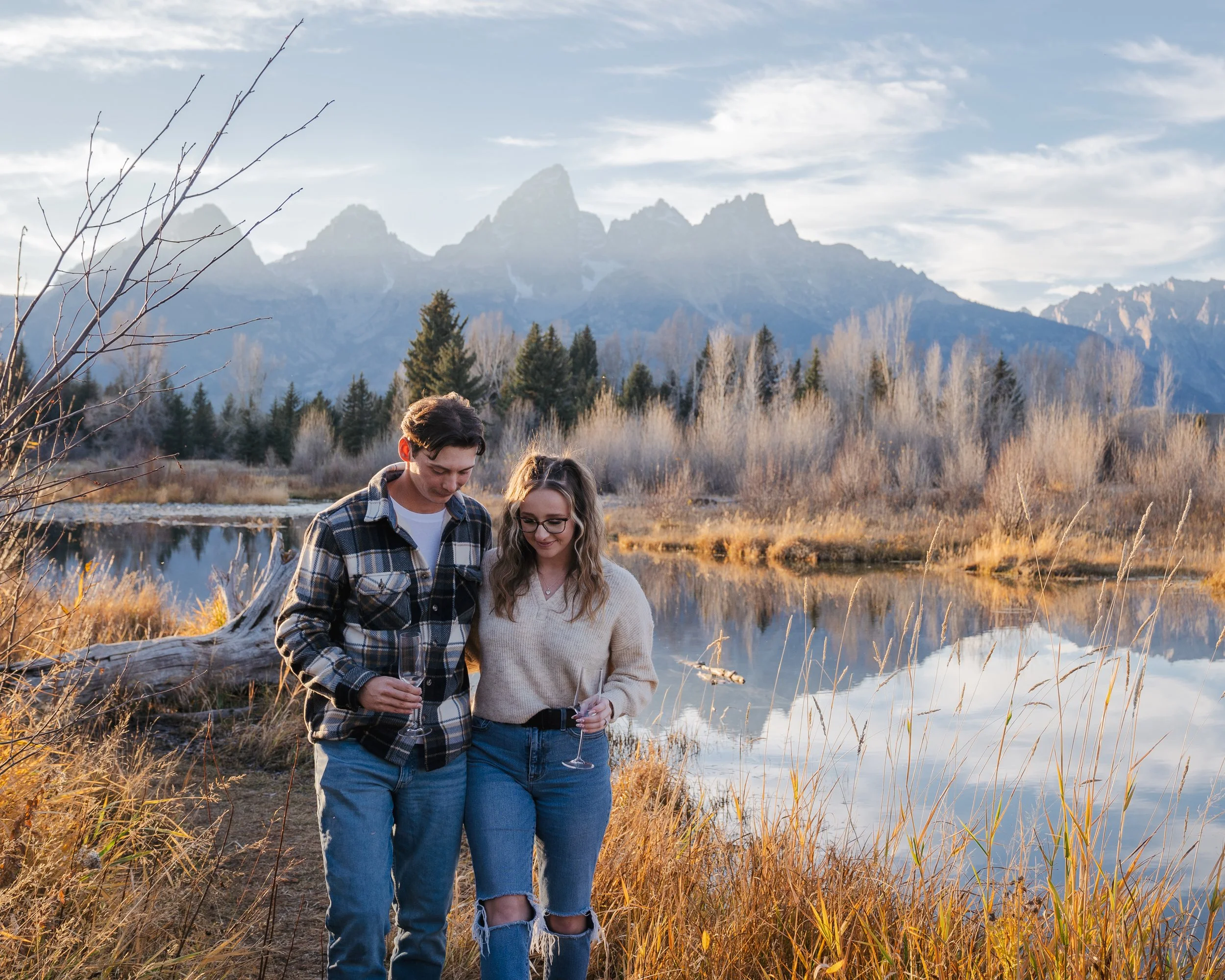 A young couple walking near a lake with mountains in the background, holding glasses of champagne, during sunset.