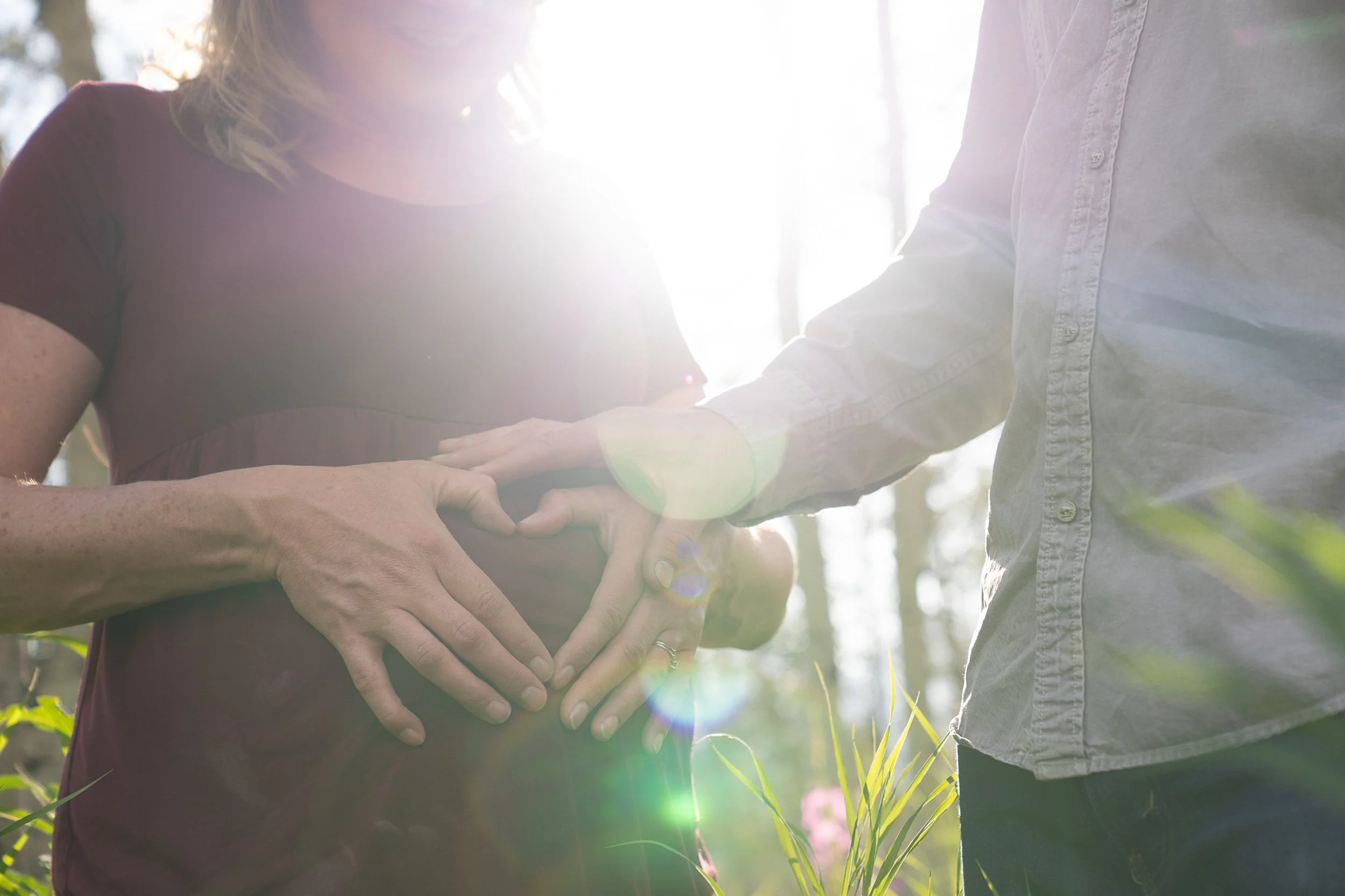 A pregnant woman and her partner are standing outdoors, forming a heart shape with their hands over her baby bump, with sunlight shining through the trees in the background.