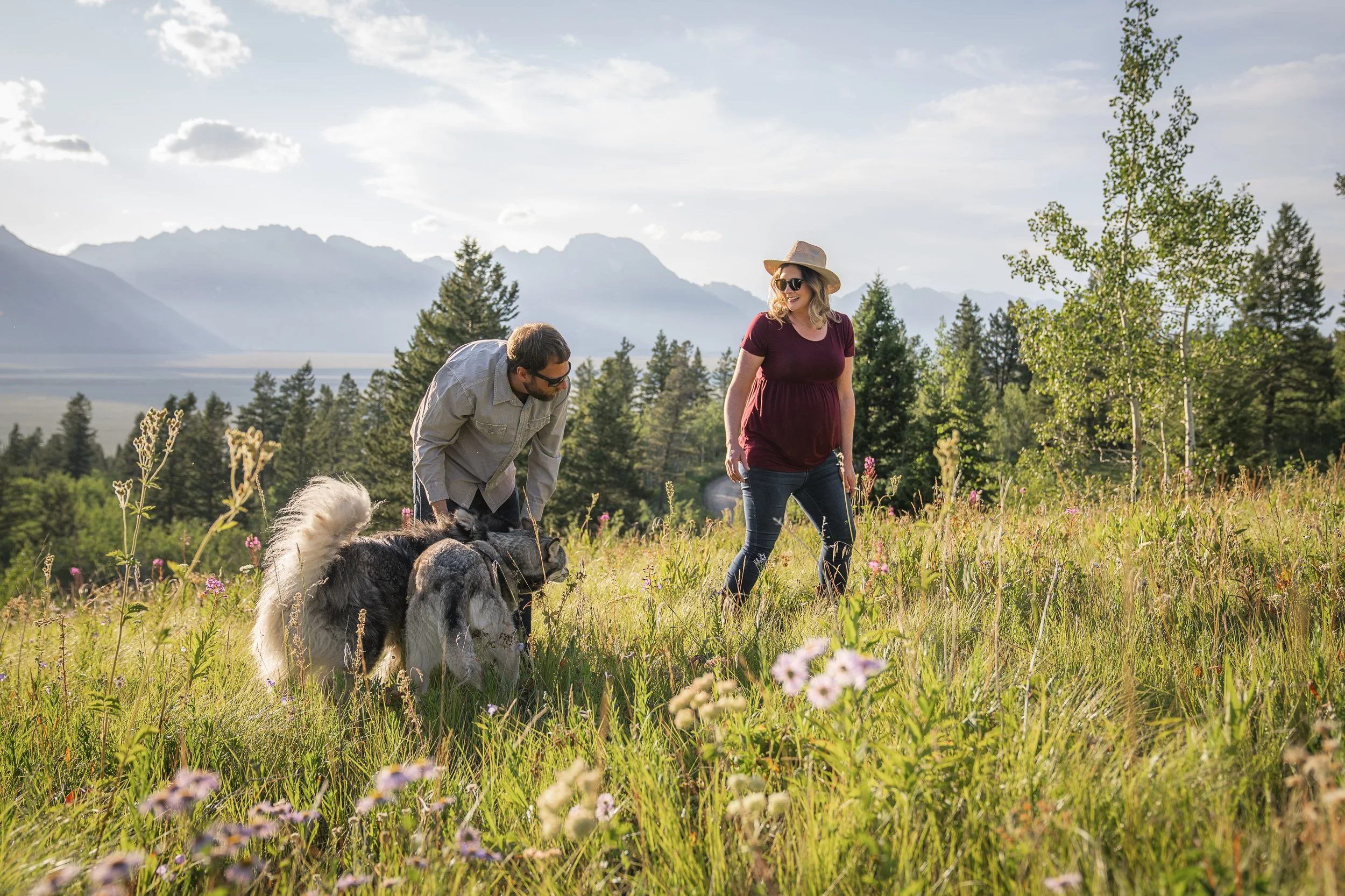 A man and woman with a dog walking in a grassy field with wildflowers, trees, mountains, and a cloudy sky in the background.