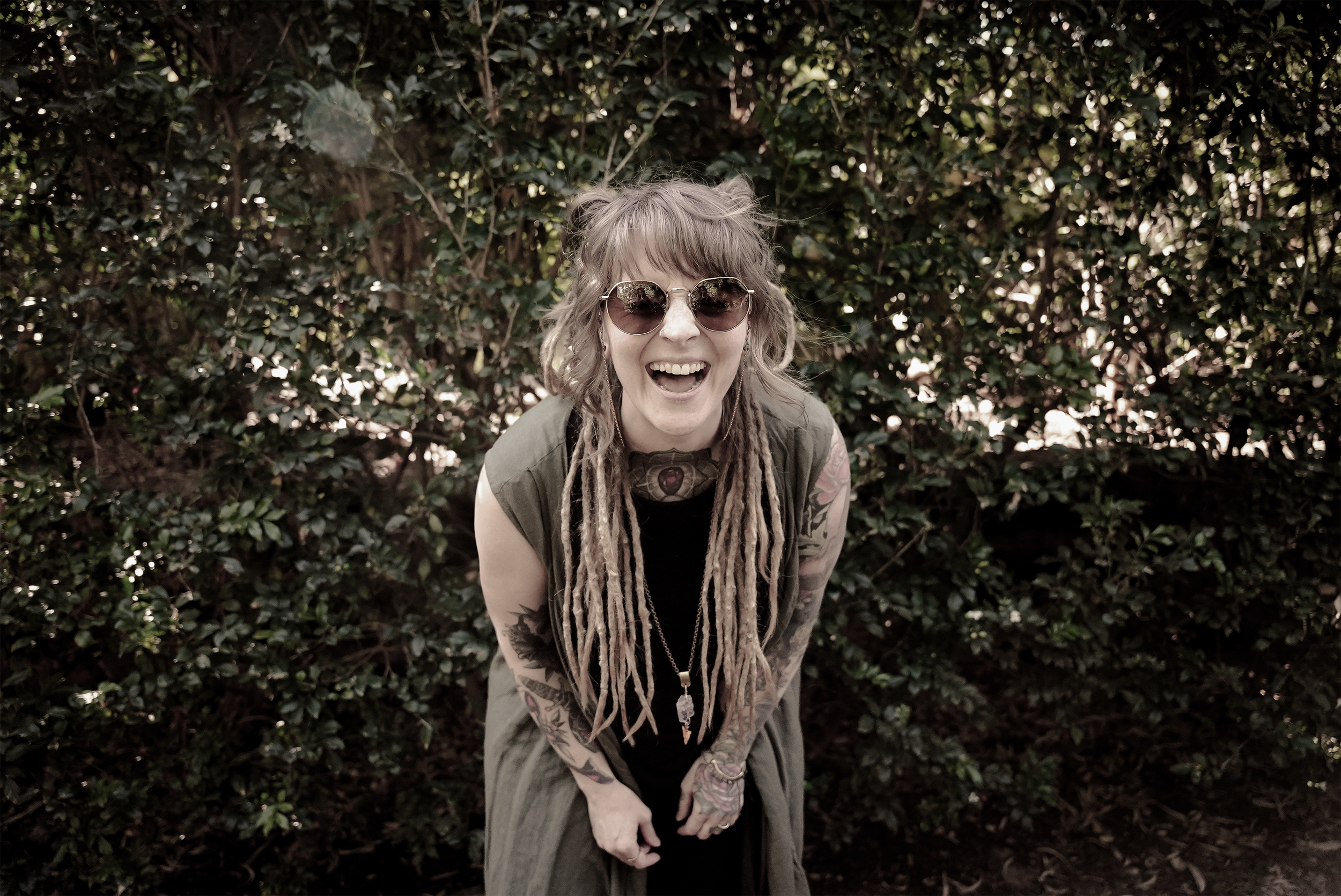 Rebecca, the face behind Reblou Design, with dreadlocks, wearing sunglasses and a sleeveless jacket, smiling and laughing outdoors in front of green bushes.