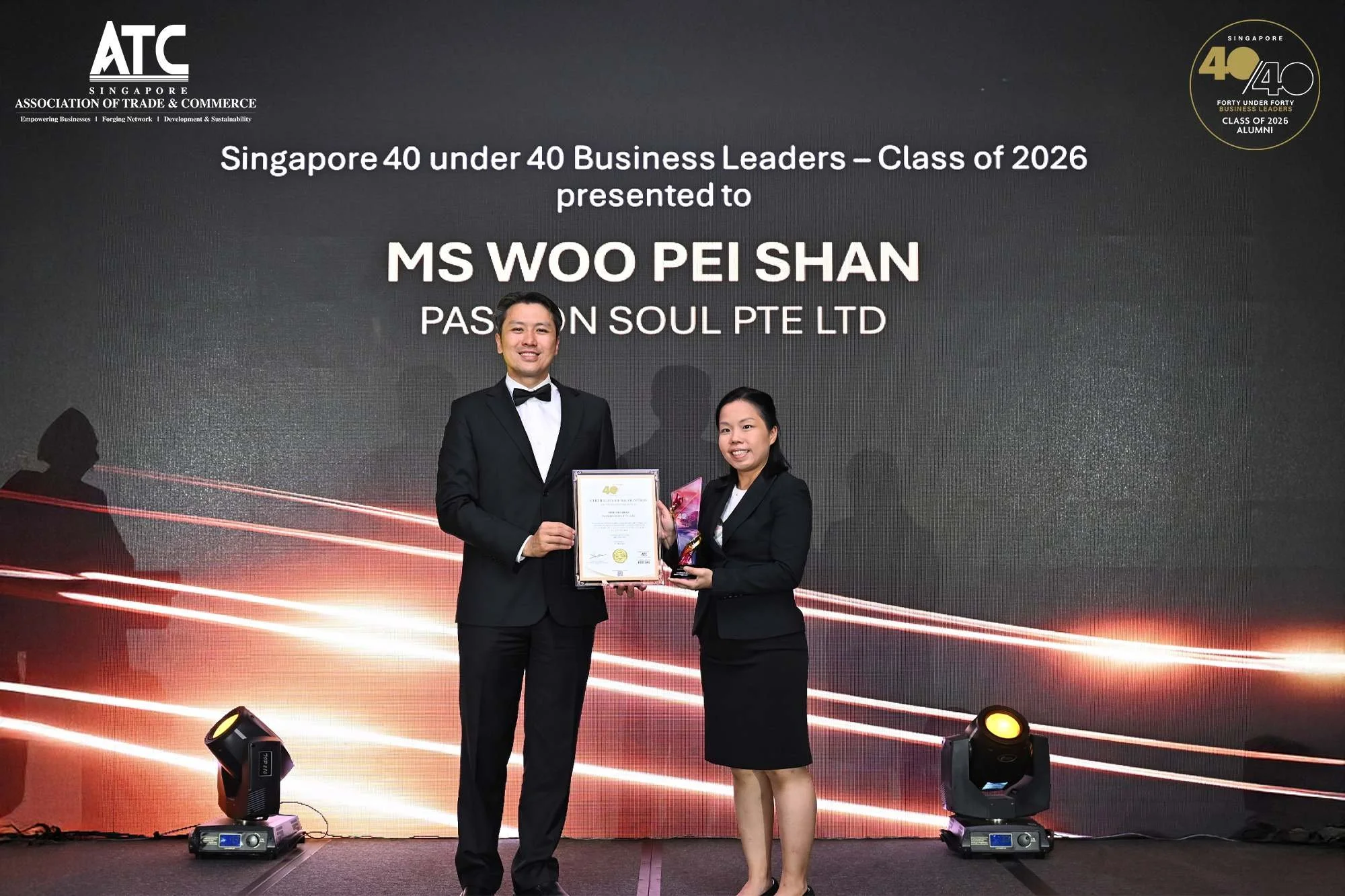 Two women in formal attire standing on stage, one presenting an award to the other, in front of a screen displaying the event name 'Singapore 40 under 40 Business Leaders, Class of 2026,' with lights and stage equipment visible.